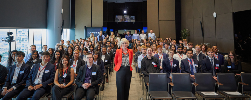 Kirsten Goodwin, a woman with short blonde hair and wearing a bright orange jacket, black and white top and black trousers, is standing smiling in front of a room full of graduates at the HSBC Global Graduate Induction.