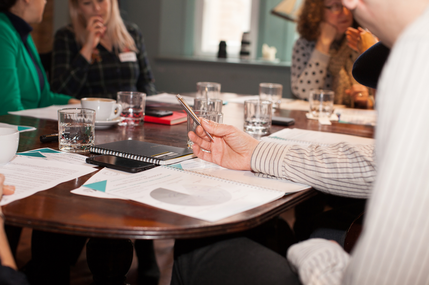 A small group gathers around a wooden table for a business meeting, with notebooks, papers featuring pie charts, water glasses, and coffee cups spread across the surface. In the foreground, a hand holds a pen while gesturing over an open notebook, with colleagues in conversation in the background.